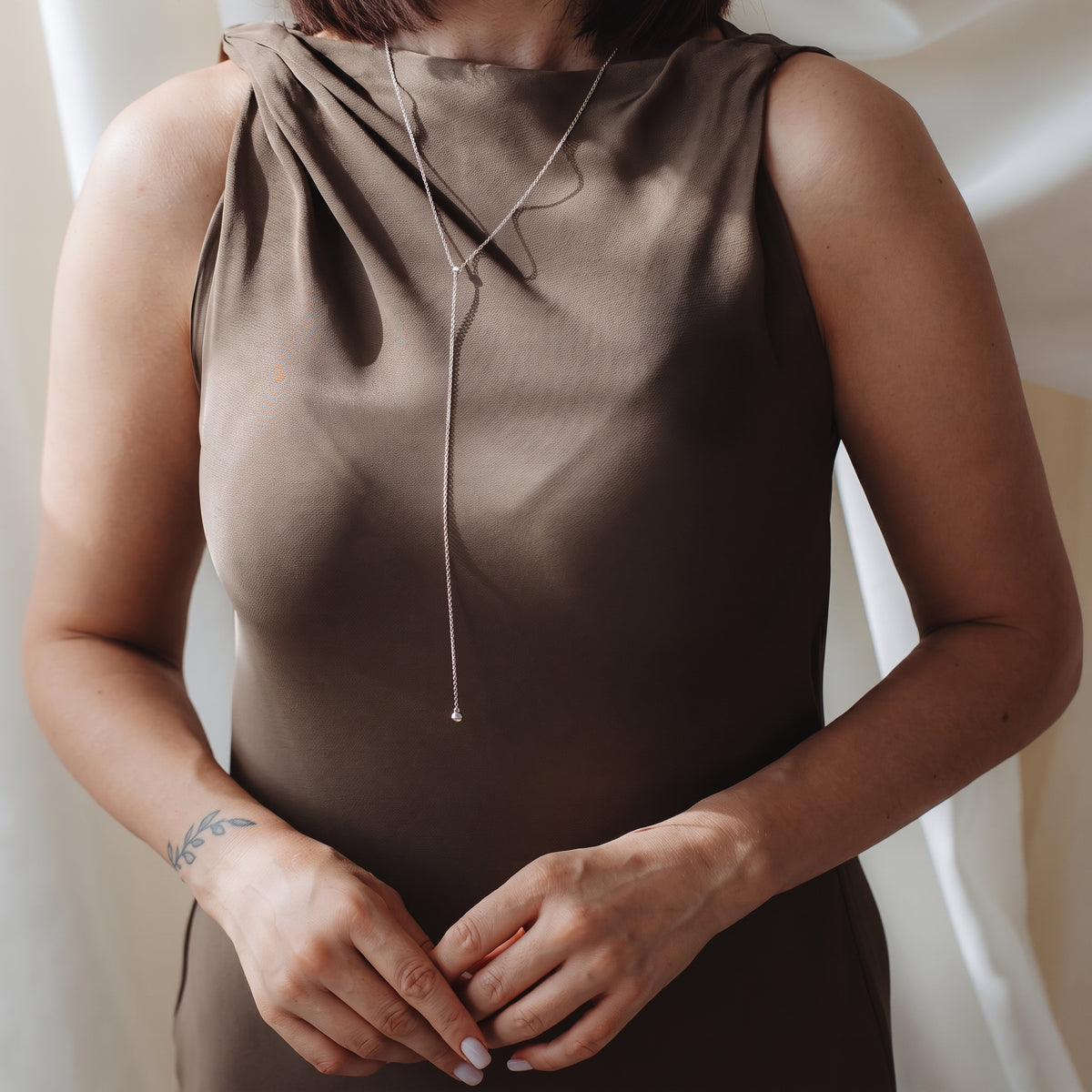 Primo piano di una persona che indossa una collana bolo (lariat) in argento sterling sopra un abito in raso color taupe, in una morbida luce interna.