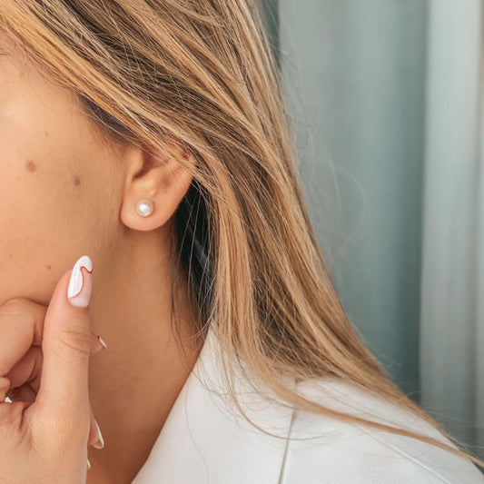 Primo piano di un orecchio femminile con un orecchino a perno in argento con perla, mentre una mano con unghie curate tocca il viso.