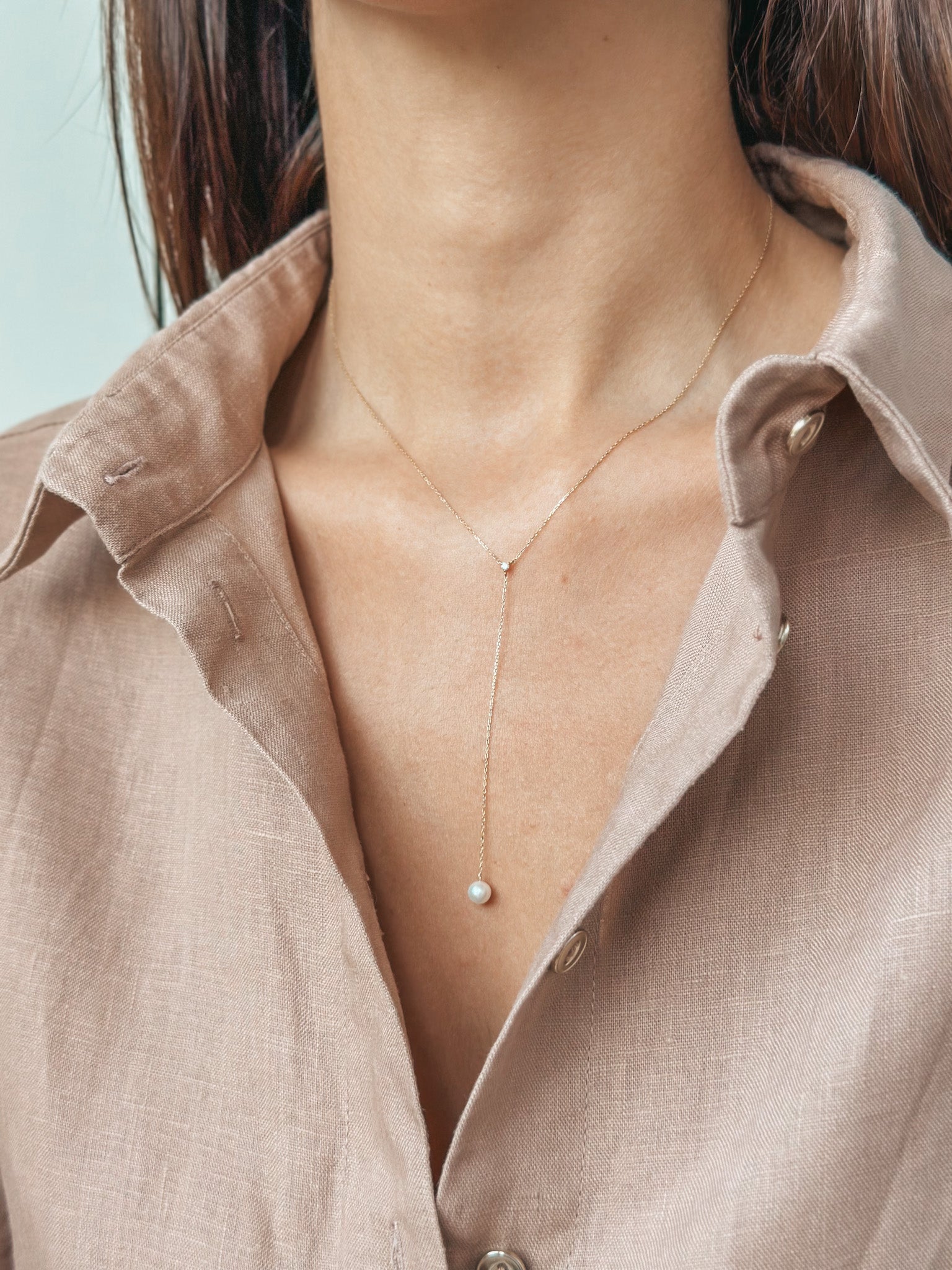 Una donna che indossa una collana d'oro con un piccolo pendente di perla, camicia beige parzialmente visibile