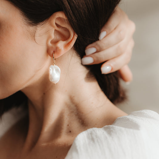 Primo piano di un orecchio che indossa un orecchino a cerchio in argento 925 placcato oro con una grande perla barocca bianca; mano con manicure neutra vicino all'orecchio in morbida luce naturale.