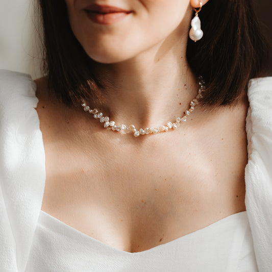Primo piano di una modella che indossa una delicata collana di perle keshi rosa e un orecchino pendente con perla barocca placcata in oro, abbinata a un abito bianco in morbida luce naturale.