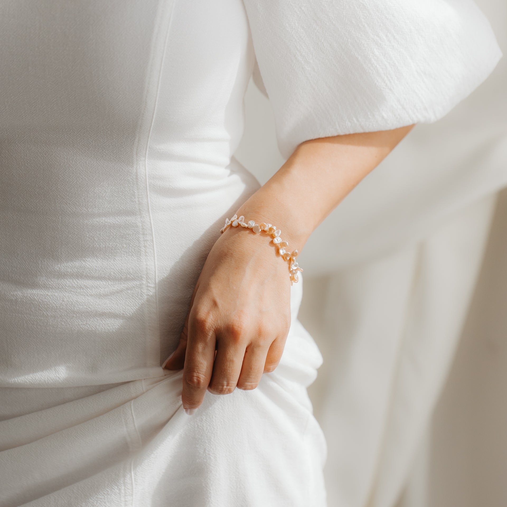 Primo piano di un bracciale in argento sterling placcato oro con perle keshi rosa al polso di una persona, abbinato a un abito bianco in luce soffusa.