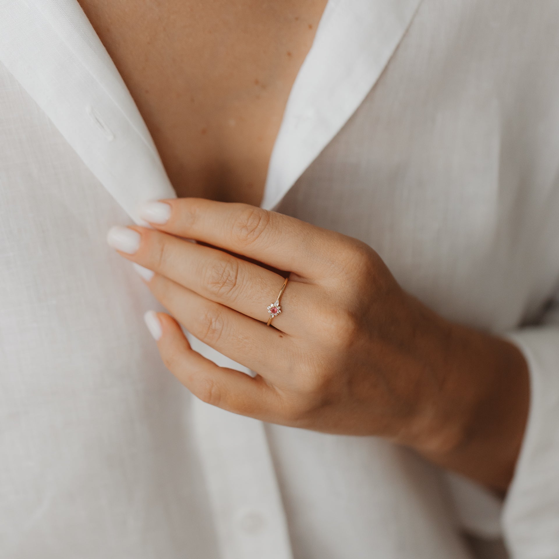 Primo piano di una mano che indossa l'anello Flora in oro giallo 14K con zaffiro bianco e tormalina rosa a grappolo floreale, sullo sfondo di una camicia di lino bianca.