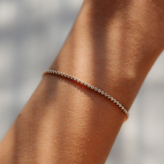 Primo piano di un delicato bracciale tennis in oro giallo 14K con piccoli diamanti rotondi indossato al polso, illuminato da una morbida luce naturale.