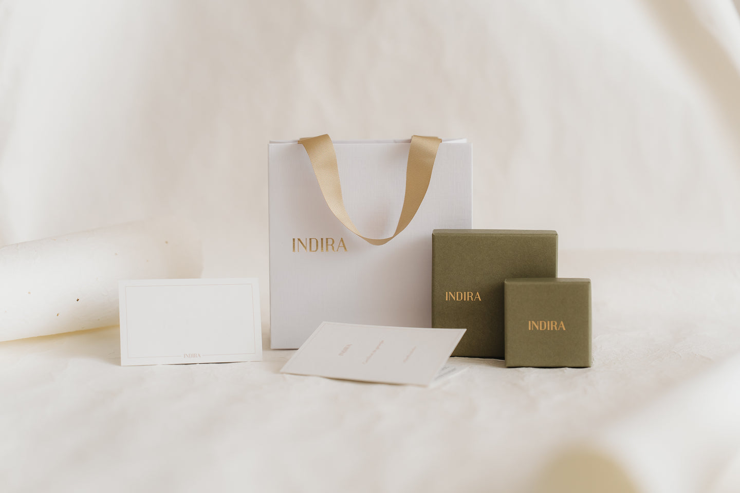 Foto in studio minimalista ed elegante del set di confezioni regalo INDIRA: una borsa bianca con manici in nastro dorato, due scatole per gioielli color oliva e biglietti vuoti su uno sfondo avorio tenue.