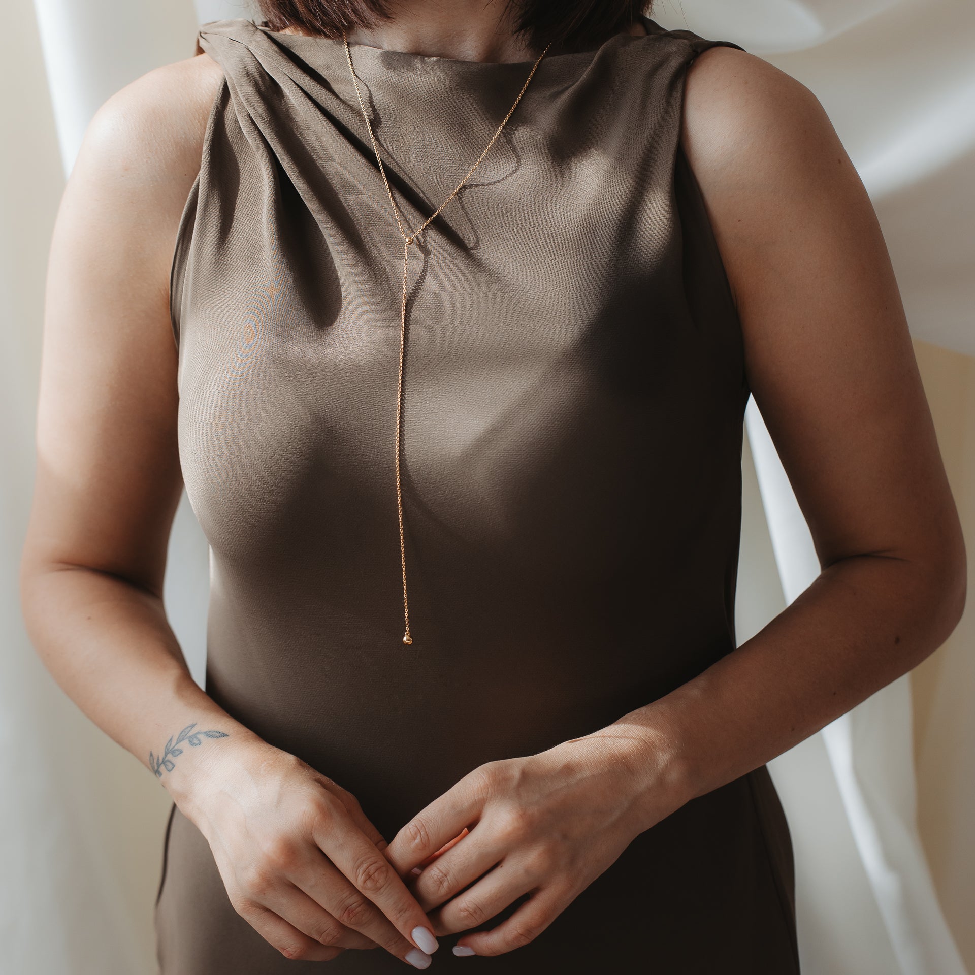 Primo piano di una persona che indossa una collana lariat con bola in argento 925 placcato oro sopra un abito di raso marrone, con le mani delicatamente congiunte.