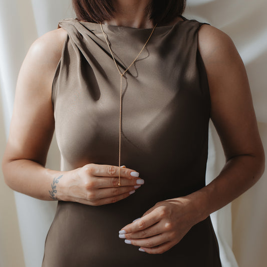 Primo piano di una persona che indossa una collana lariat (Bola) in argento 925 placcato oro sopra un abito in raso taupe, con le mani che regolano delicatamente il pendente a goccia.