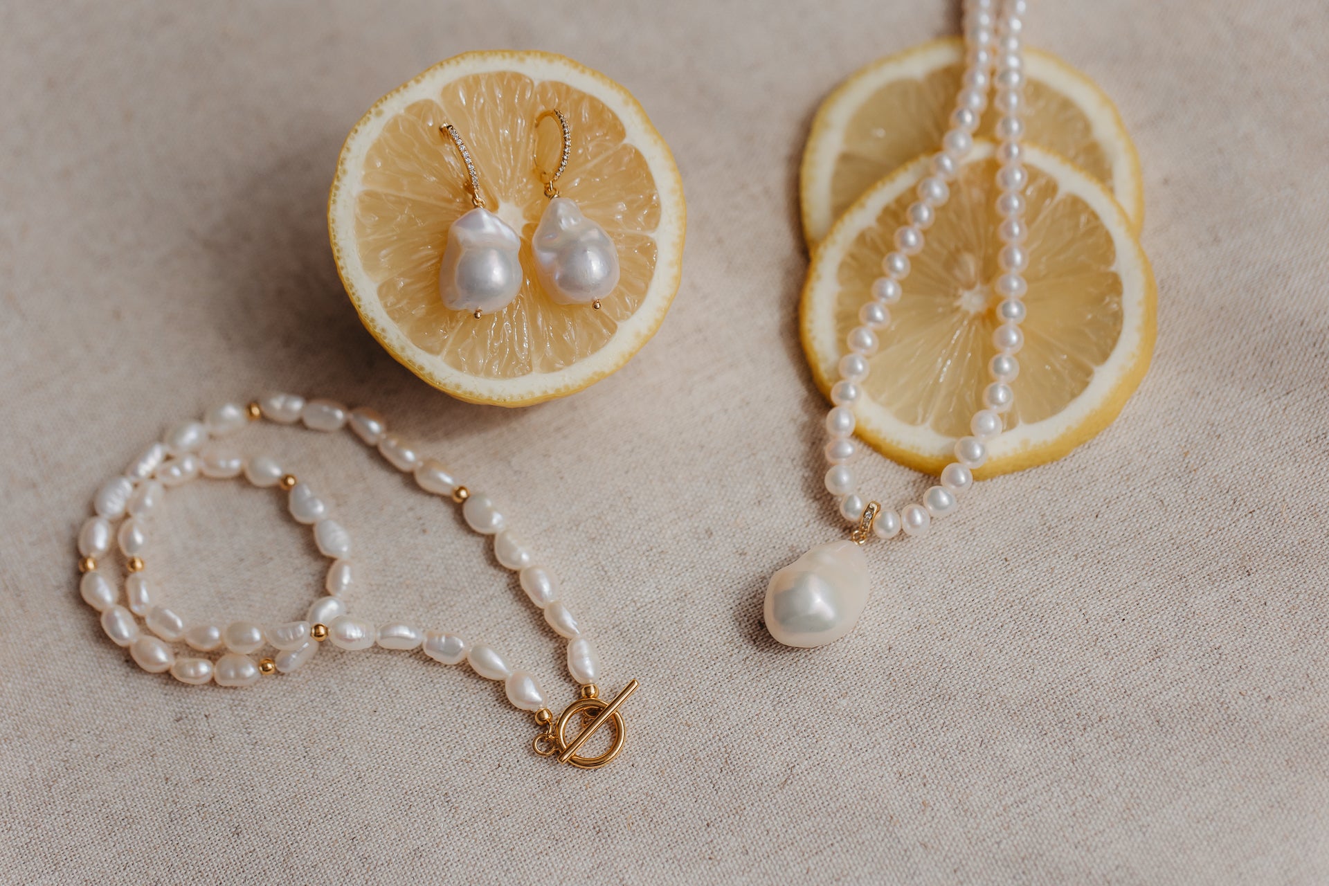 Orecchini con perle barocche su cerchi in argento sterling placcato oro, inseriti in una metà di limone, con collana e bracciale di perle coordinati su lino beige.