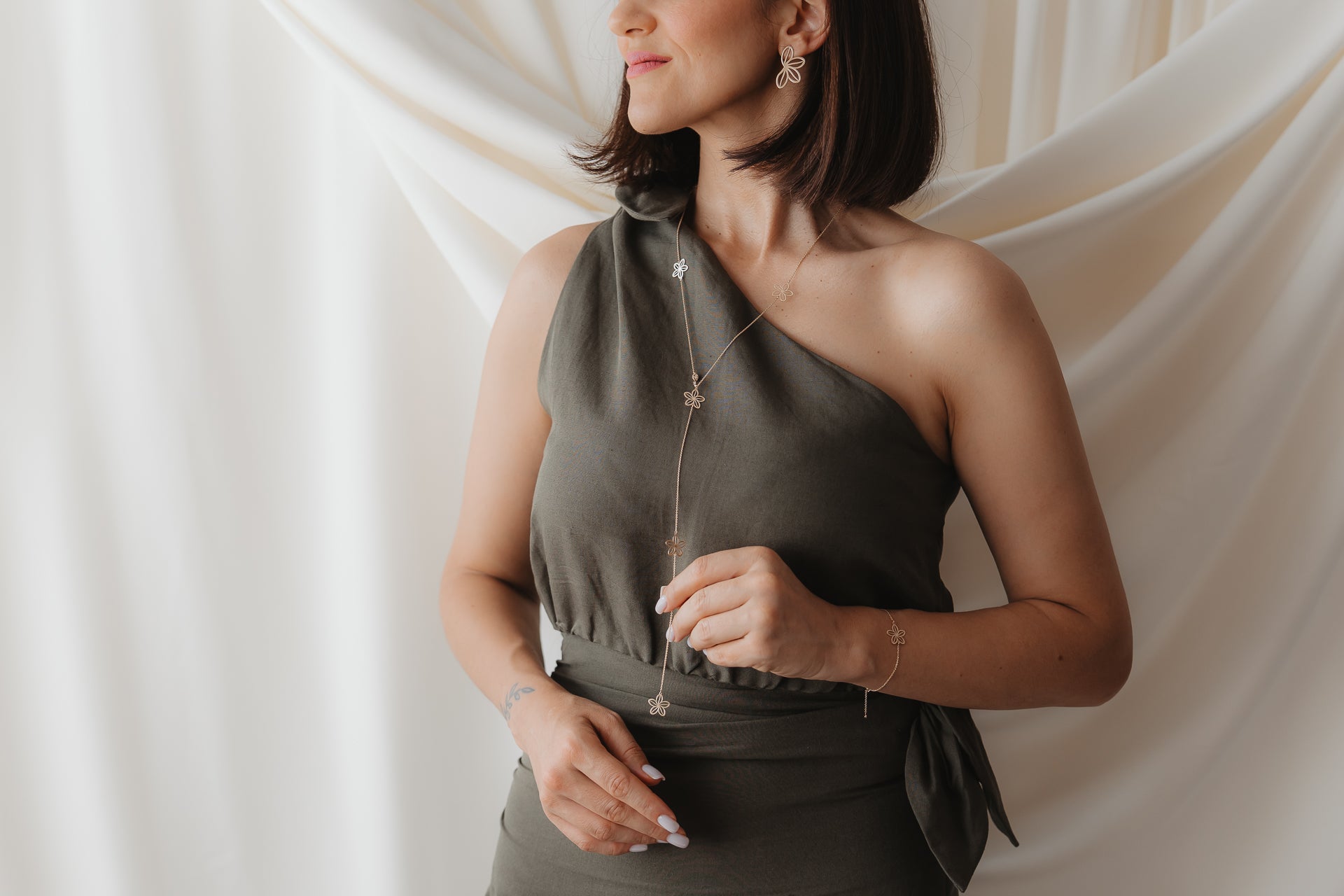 Primo piano di una modella che indossa la collana lariat Wildflower Maxi in oro giallo 14K con delicati ciondoli a forma di fiore, abbinata a un abito monospalla verde oliva su uno sfondo crema morbido e drappeggiato.