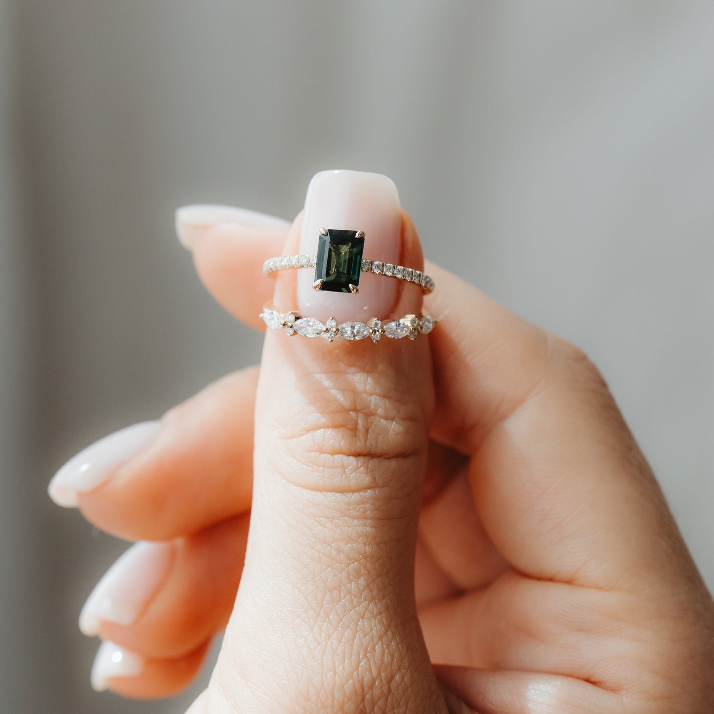 Primo piano di una mano che tiene due anelli in oro giallo: un anello Elysian con zaffiro verde taglio smeraldo e fascia con pavé di diamanti, indossato sopra una fedina Fiorelle con diamanti taglio marquise, su uno sfondo grigio tenue.