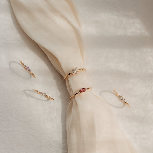 Cinque delicati anelli in oro giallo 14k con diamanti e gemme rosa, disposti su un tessuto di seta color crema annodato alla luce solare soffusa.