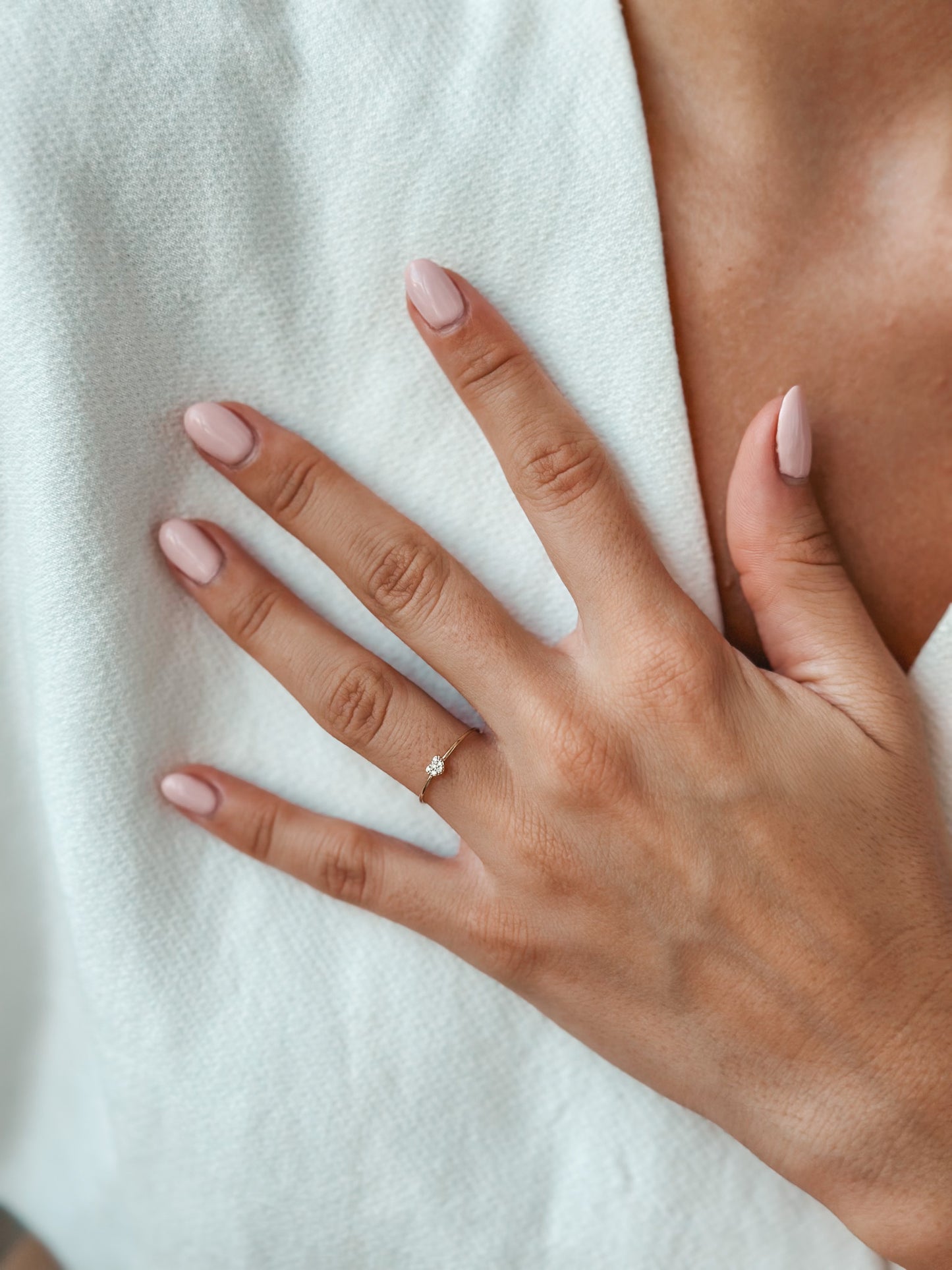 Close-up of a hand with pink polished nails wearing a delicate gold ring with a small diamond against a soft white fabric background.