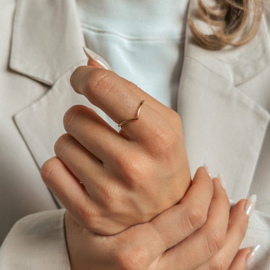 Primo piano di una mano che indossa un semplice anello in argento placcato oro a forma di V, con abbigliamento dai toni neutri sullo sfondo.