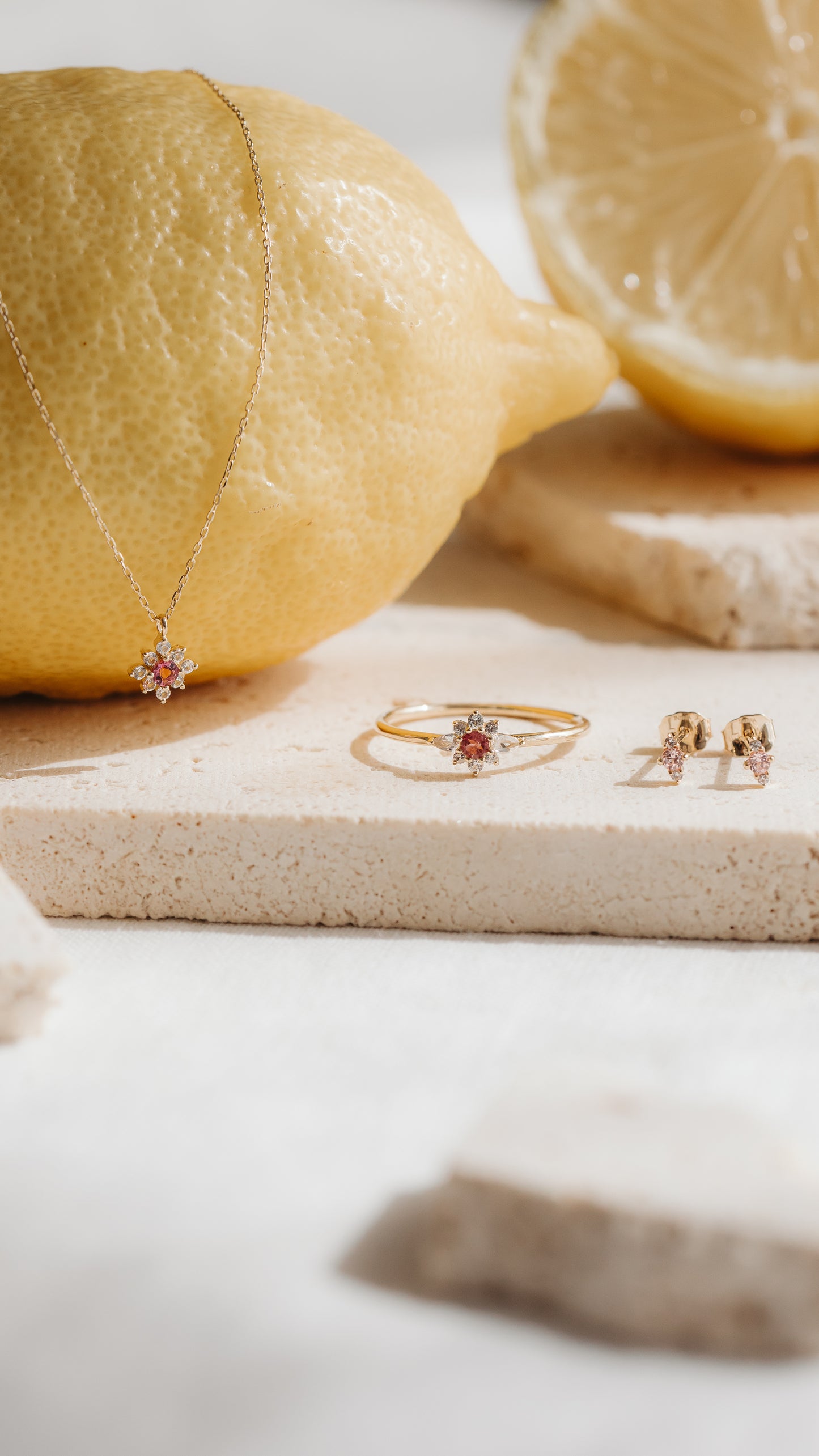 Orecchini a bottone Flora in oro giallo 14K con morganite rosa e zaffiri bianchi, accostati a un anello e a un ciondolo coordinati su piastrelle in pietra con limoni.