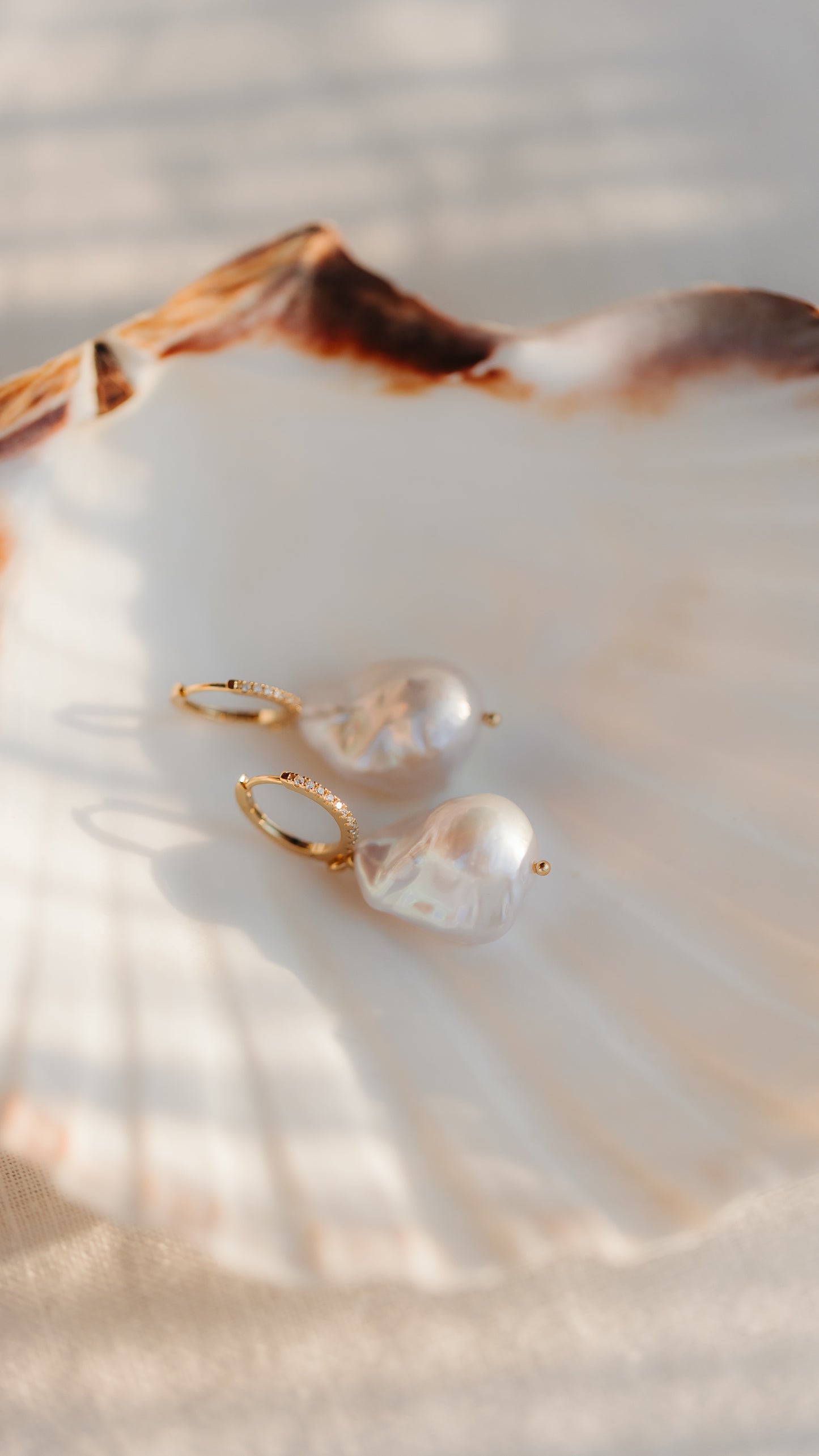 Orecchini a cerchio in argento 925 placcato oro con grandi perle barocche, adagiati su una conchiglia di capesanta in una calda luce naturale.