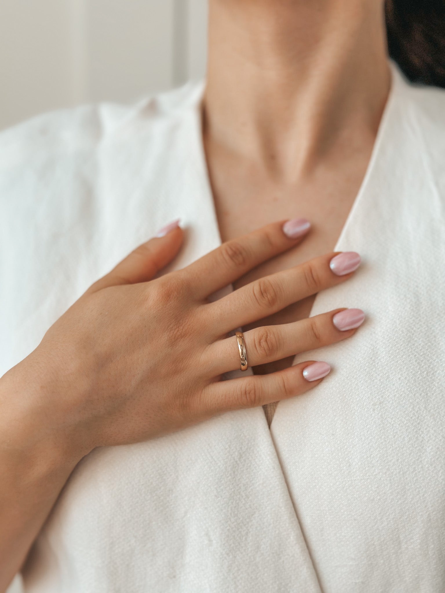 Close-up of a hand with a pink-polished manicure resting on the chest, wearing a gold wedding band and a white textured garment.
