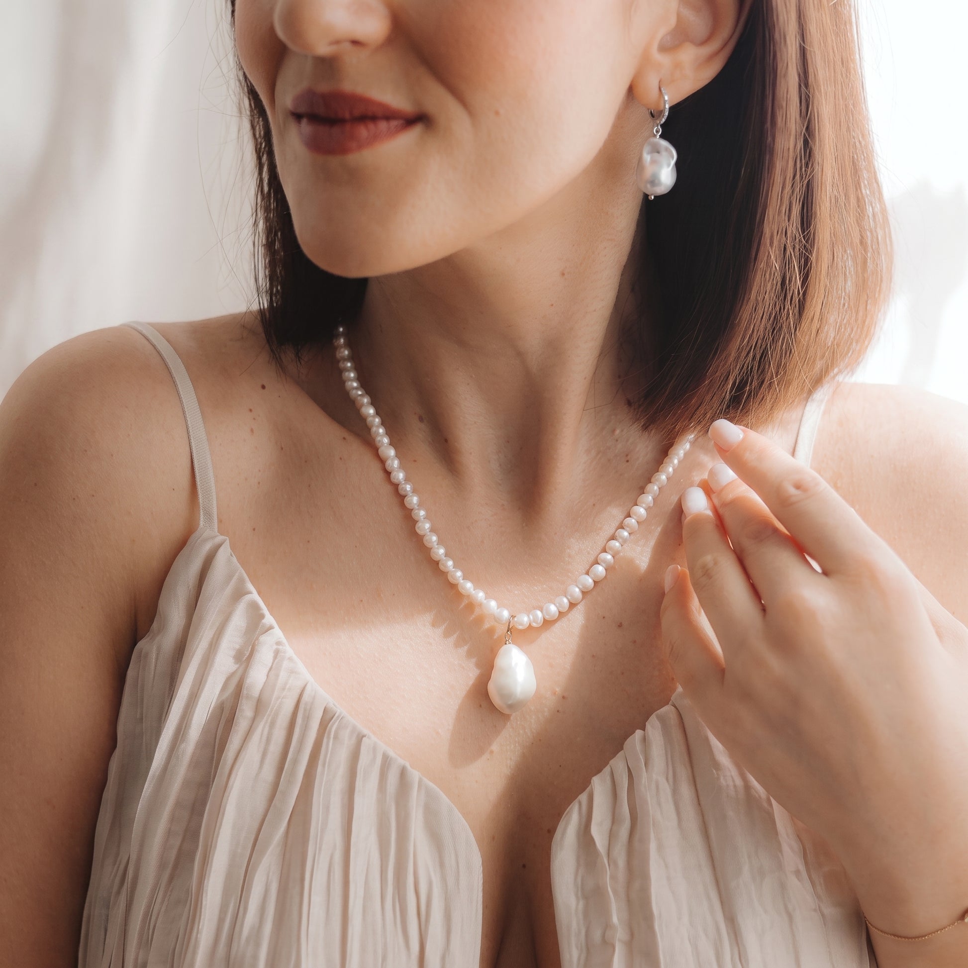Primo piano di una modella che indossa un elegante orecchino pendente con perla barocca in argento 925 rodiato, con un piccolo dettaglio in zirconia, e una collana di perle coordinata; la mano è vicino alla clavicola, in una morbida luce naturale.