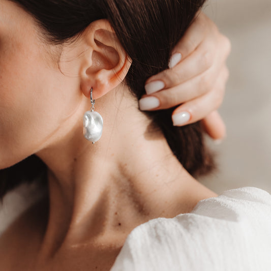 Primo piano di un orecchio con orecchino a cerchio in argento 925 rodiato con una grande perla barocca pendente, con una mano che tocca i capelli.