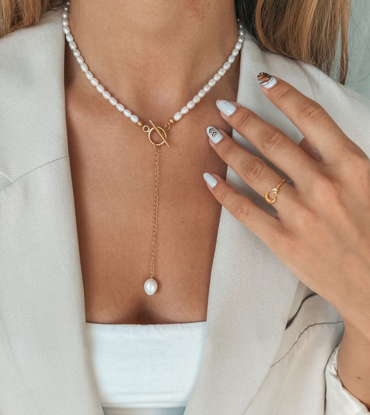 Primo piano del collo di una donna ornato con una collana di perle con catena d'oro e una perla pendente, completata da un anello d'oro minimalista sul dito.