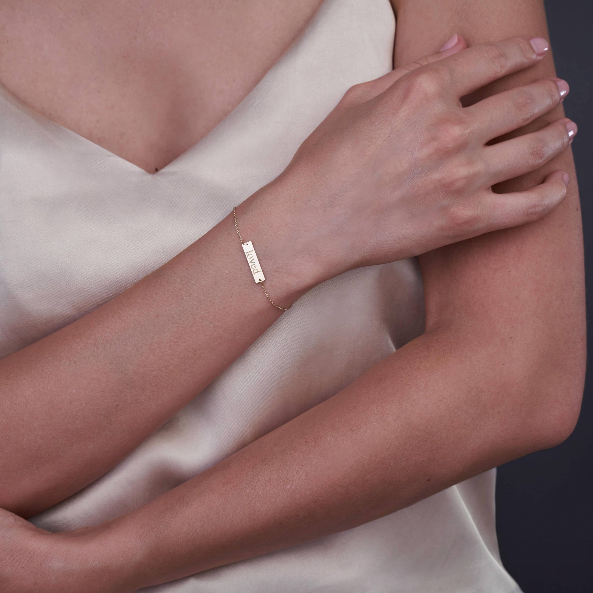 Primo piano di una persona che indossa un braccialetto in oro 14K con la scritta 'Loved' incisa, su uno sfondo neutro.