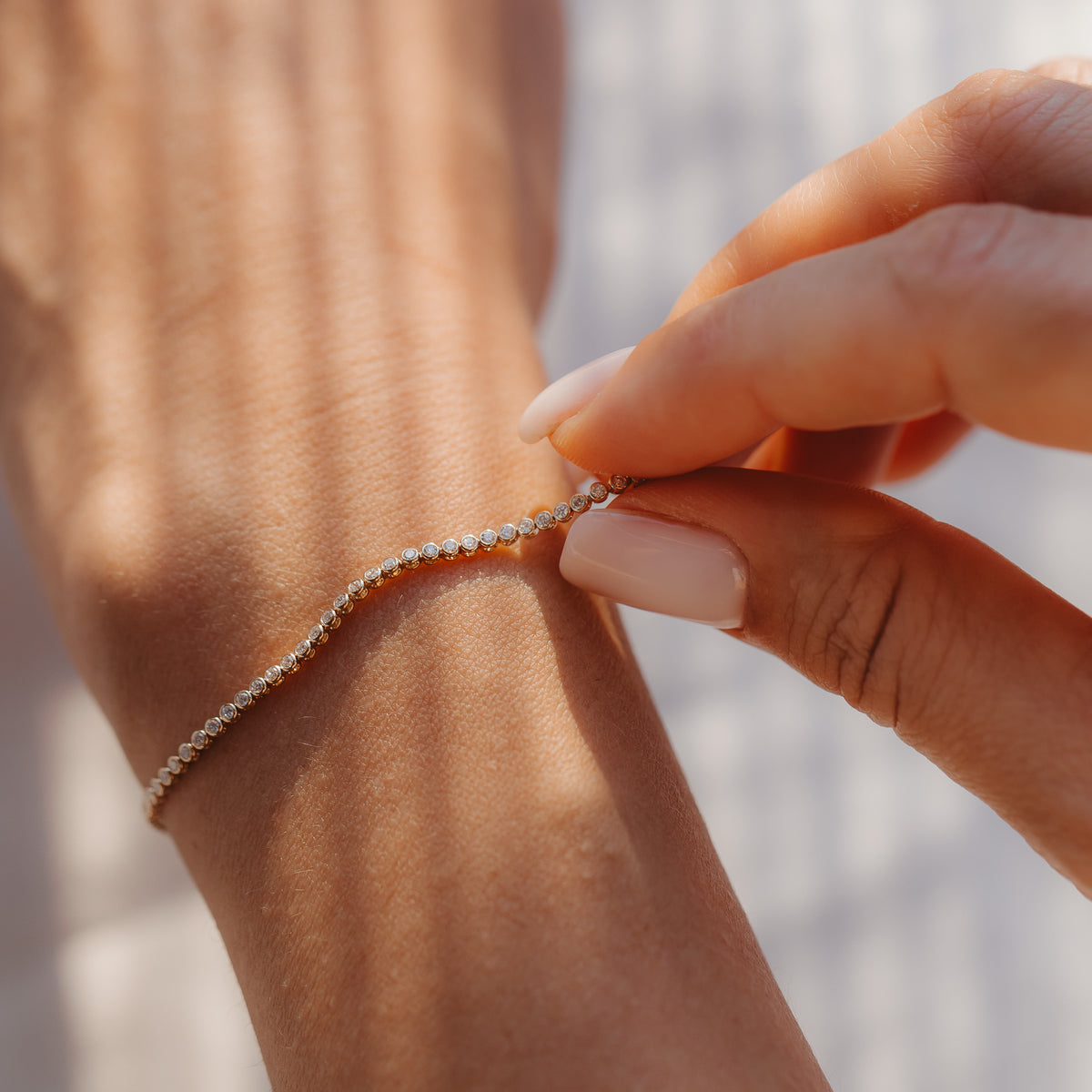 Primo piano di una mano che allaccia un elegante bracciale tennis con diamanti in oro giallo 14K a un polso, sotto una luce solare soffusa.