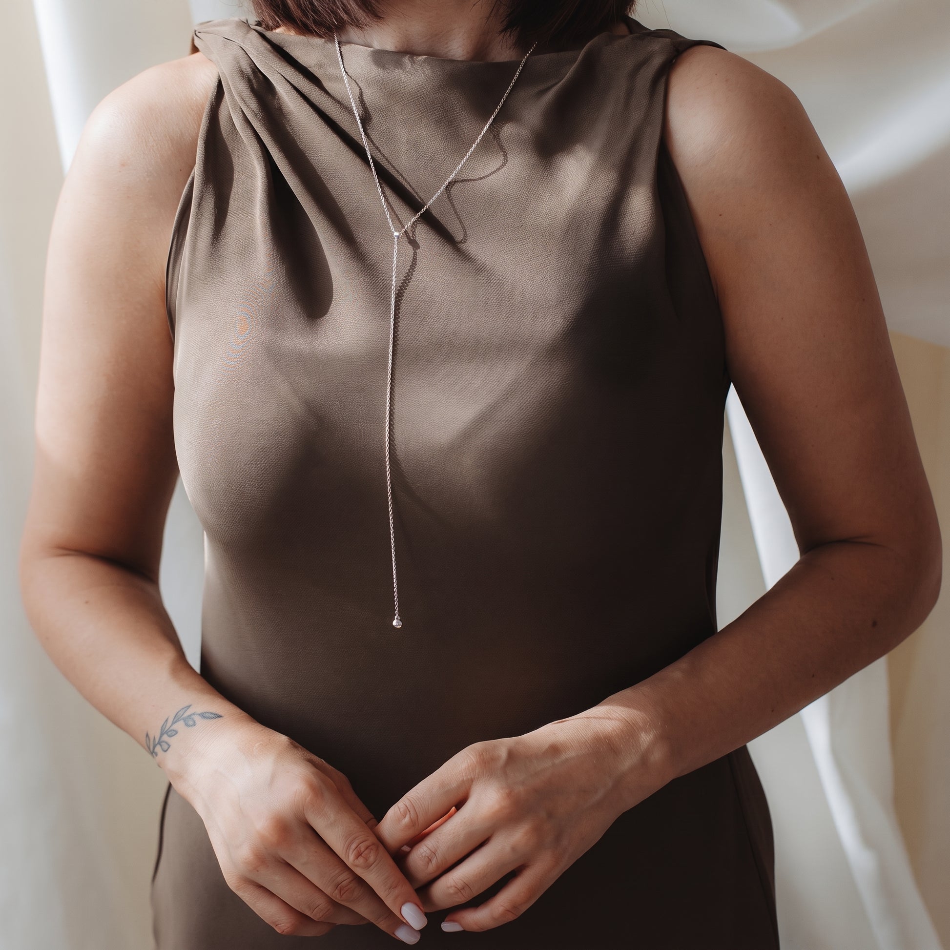 Primo piano di una persona che indossa una collana bolo (lariat) in argento sterling sopra un abito in raso color taupe, in una morbida luce interna.