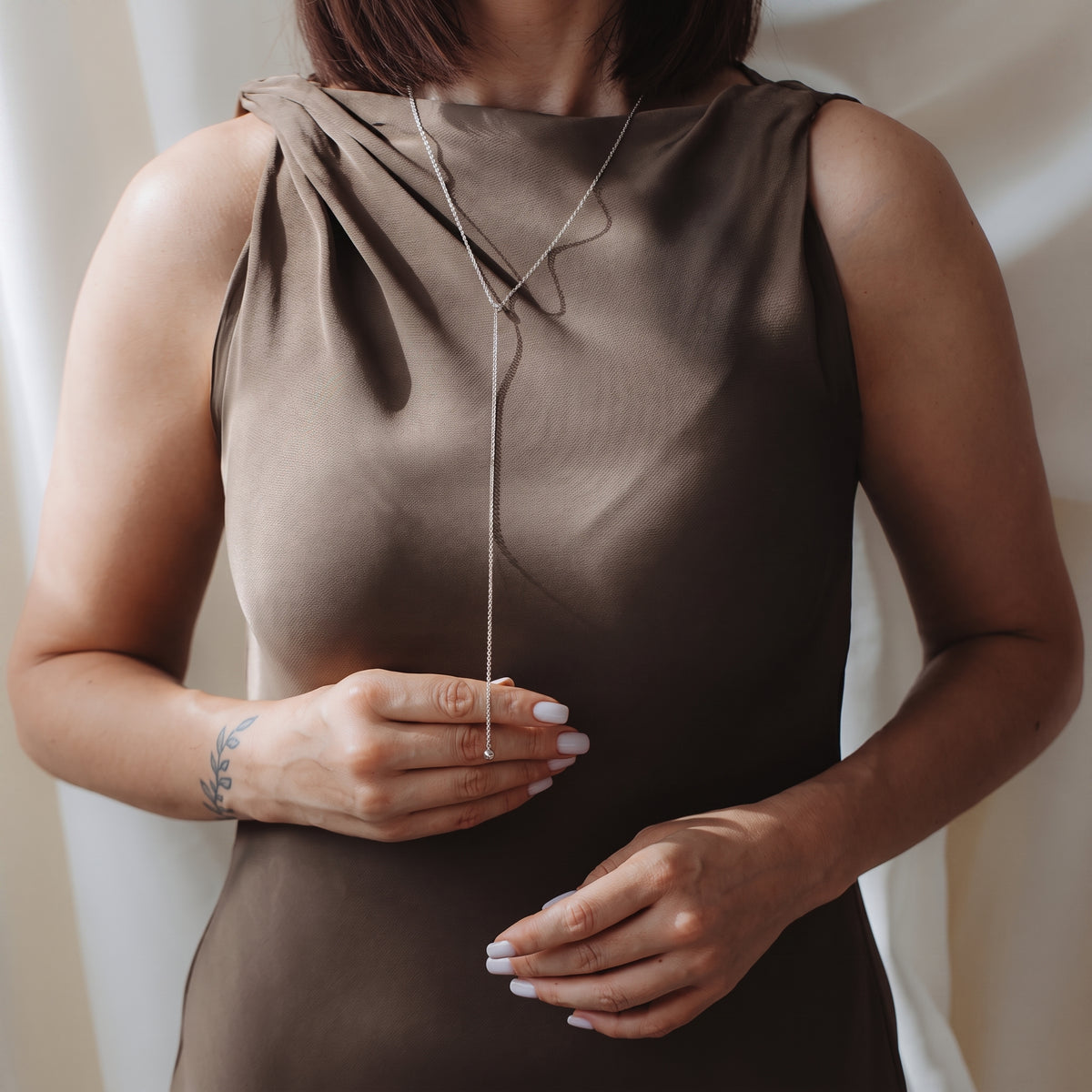Primo piano di una modella che indossa una collana lariat con bola in argento 925 sopra un abito tortora senza maniche, mentre tiene delicatamente la catenina pendente.