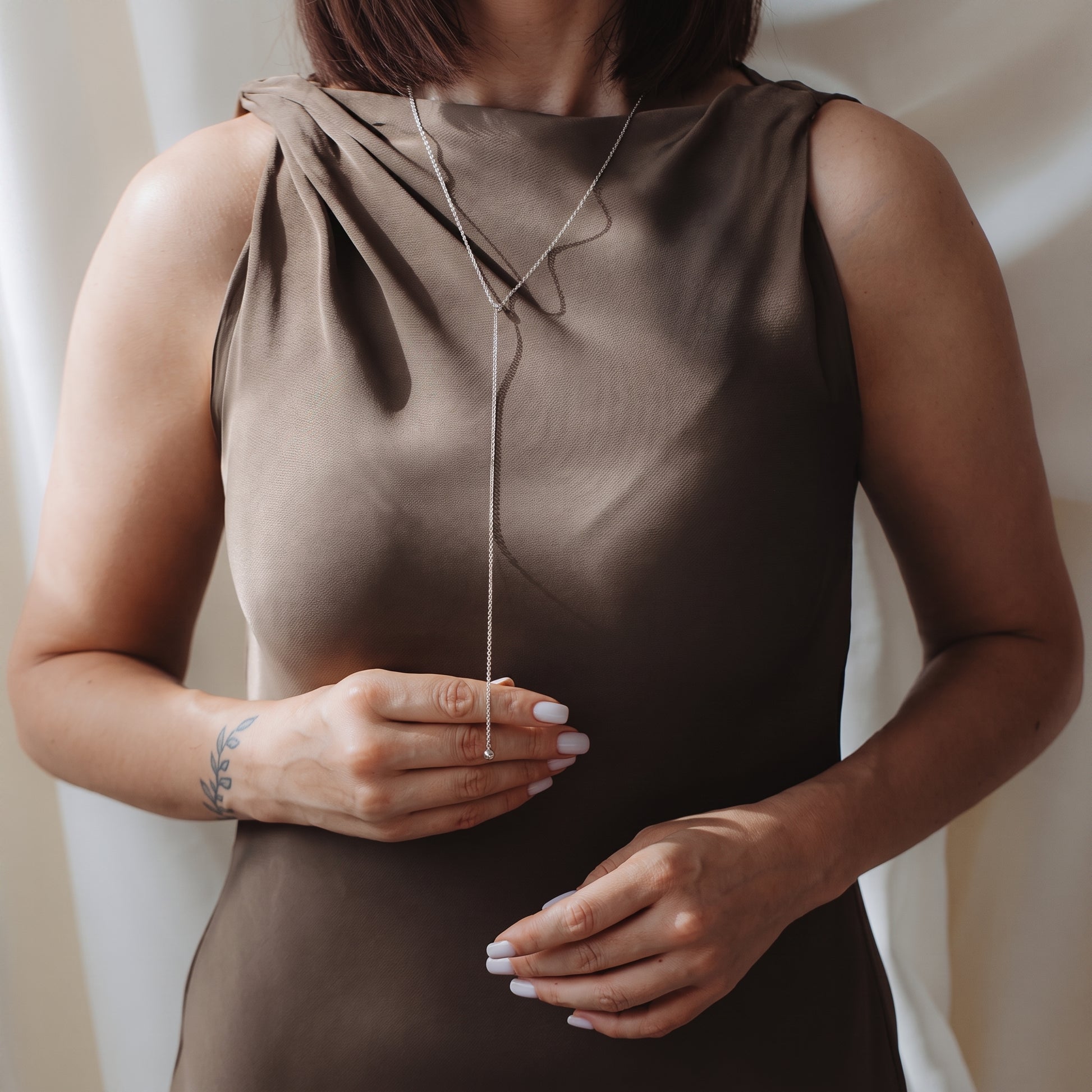 Primo piano di una modella che indossa una collana lariat con bola in argento 925 sopra un abito tortora senza maniche, mentre tiene delicatamente la catenina pendente.