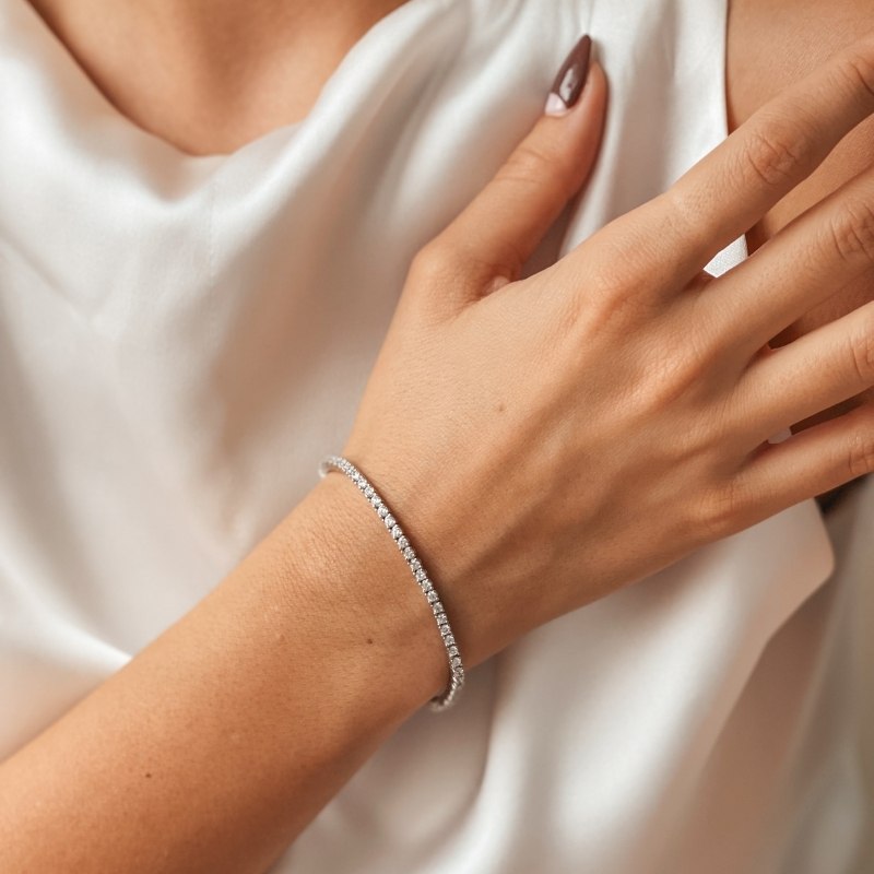 Primo piano di un polso con un bracciale tennis in argento ornato con pietre di zirconia, in contrasto con un tessuto di raso bianco.