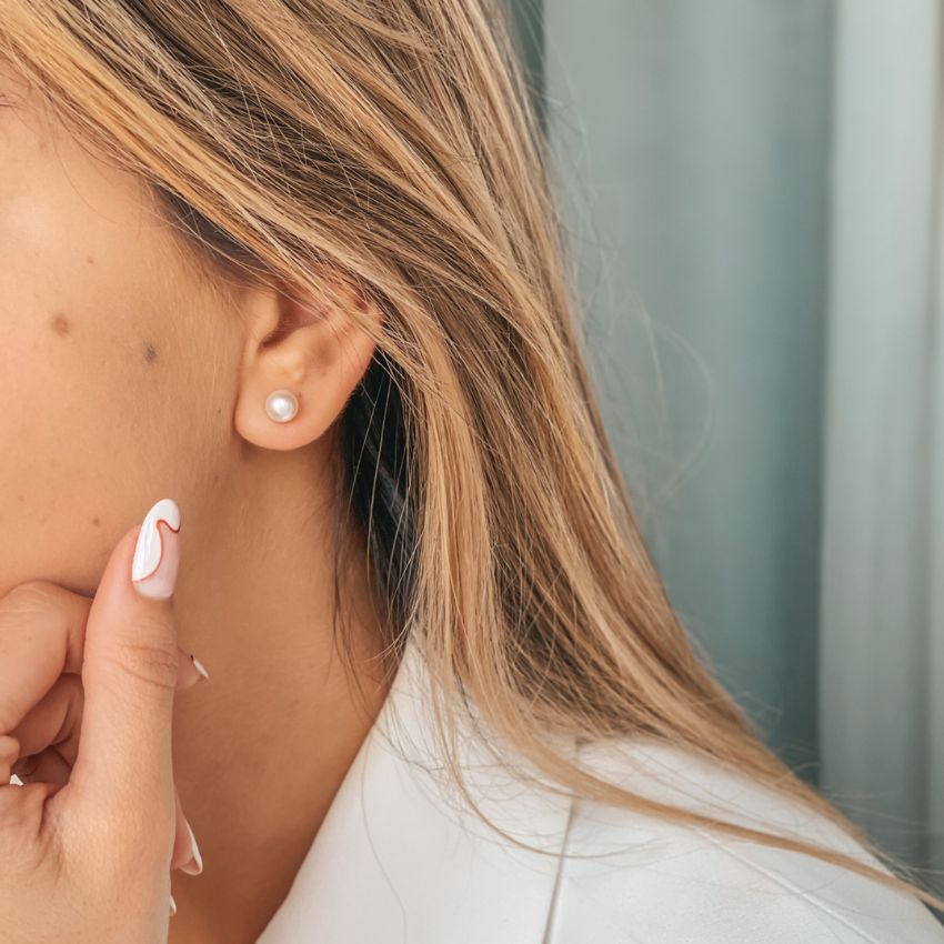Primo piano di un orecchio femminile con un orecchino a perno in argento con perla, mentre una mano con unghie curate tocca il viso.