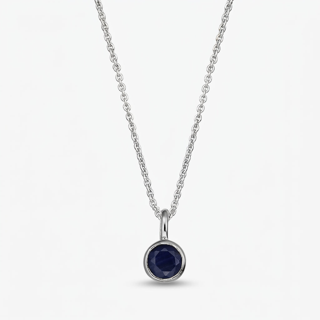 Collana in argento sterling con pietra di nascita, piccolo pendente rotondo in zaffiro blu scuro con montatura a castone su catena sottile, su sfondo bianco.