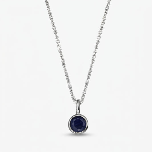 Collana in argento sterling con pietra di nascita, piccolo pendente rotondo in zaffiro blu scuro con montatura a castone su catena sottile, su sfondo bianco.