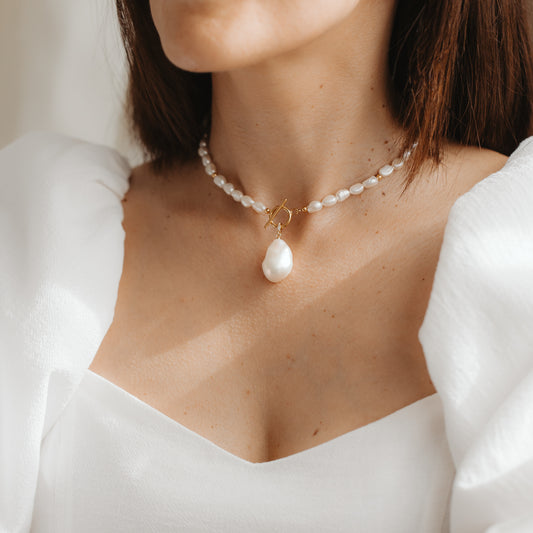 Primo piano di una donna che indossa una collana corta di perle naturali con pendente in perla barocca e chiusura a T in oro, abbinata a un elegante abito bianco.