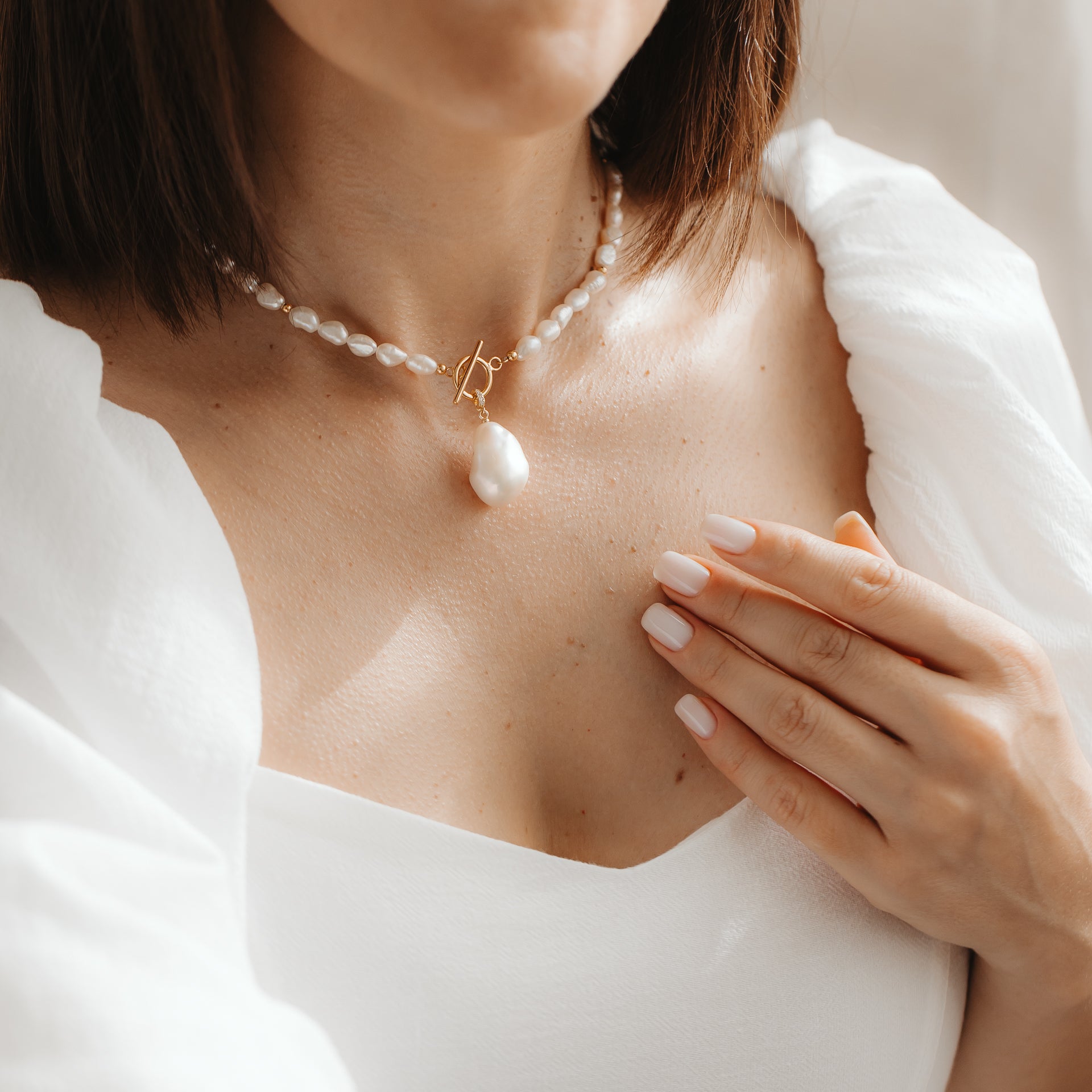 Primo piano di una donna che indossa un elegante filo di perle naturali con un pendente in perla barocca placcato in oro, mentre tocca la clavicola alla luce naturale soffusa.