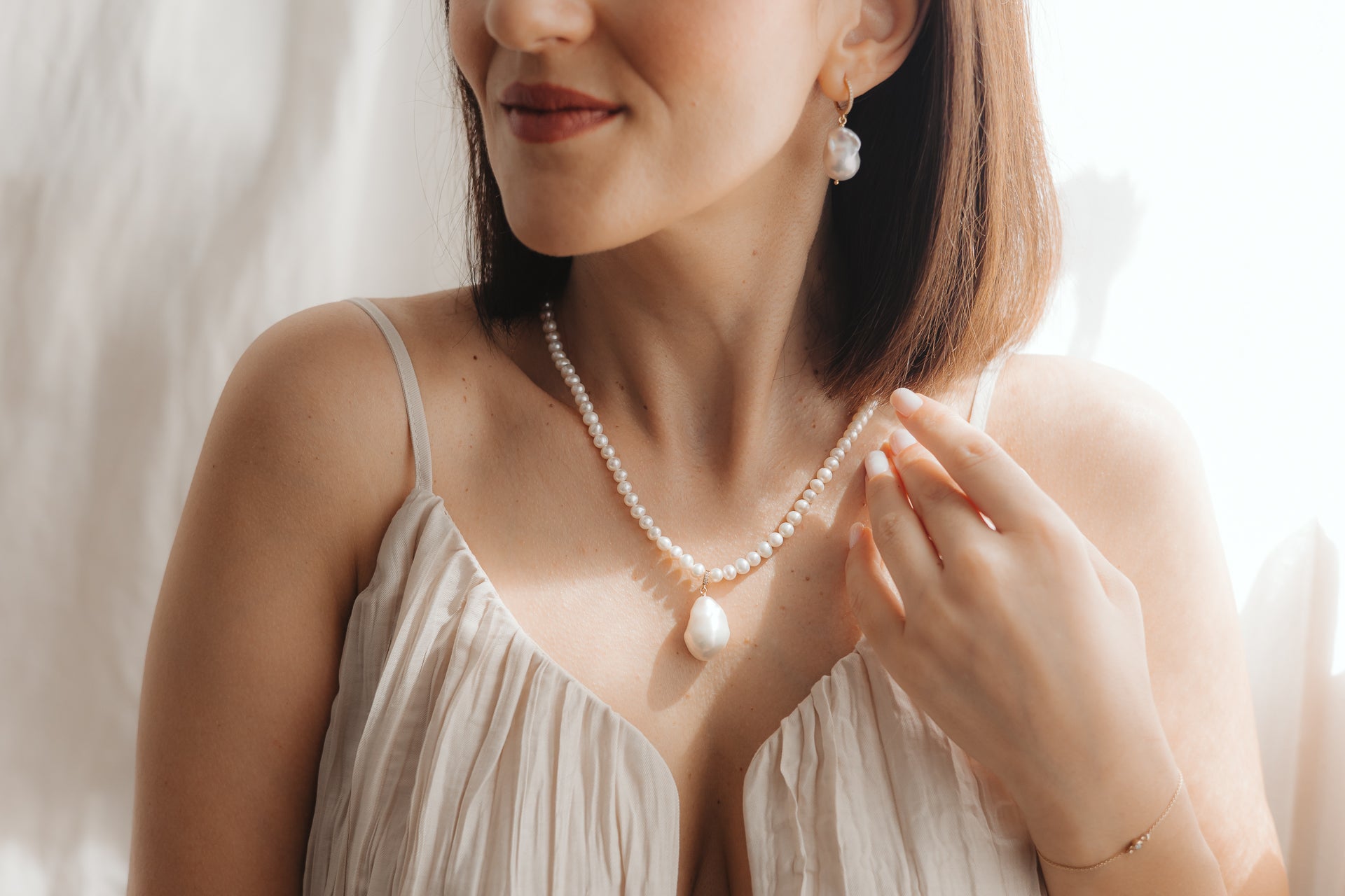 Primo piano di una donna che indossa un pendente con perla barocca su una collana di perle e un orecchino a goccia con perla coordinato in argento placcato oro, illuminata da una morbida luce naturale.