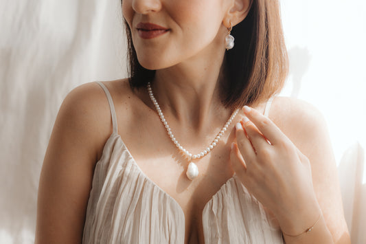Primo piano di una donna che indossa un pendente con perla barocca su una collana di perle e un orecchino a goccia con perla coordinato in argento placcato oro, illuminata da una morbida luce naturale.