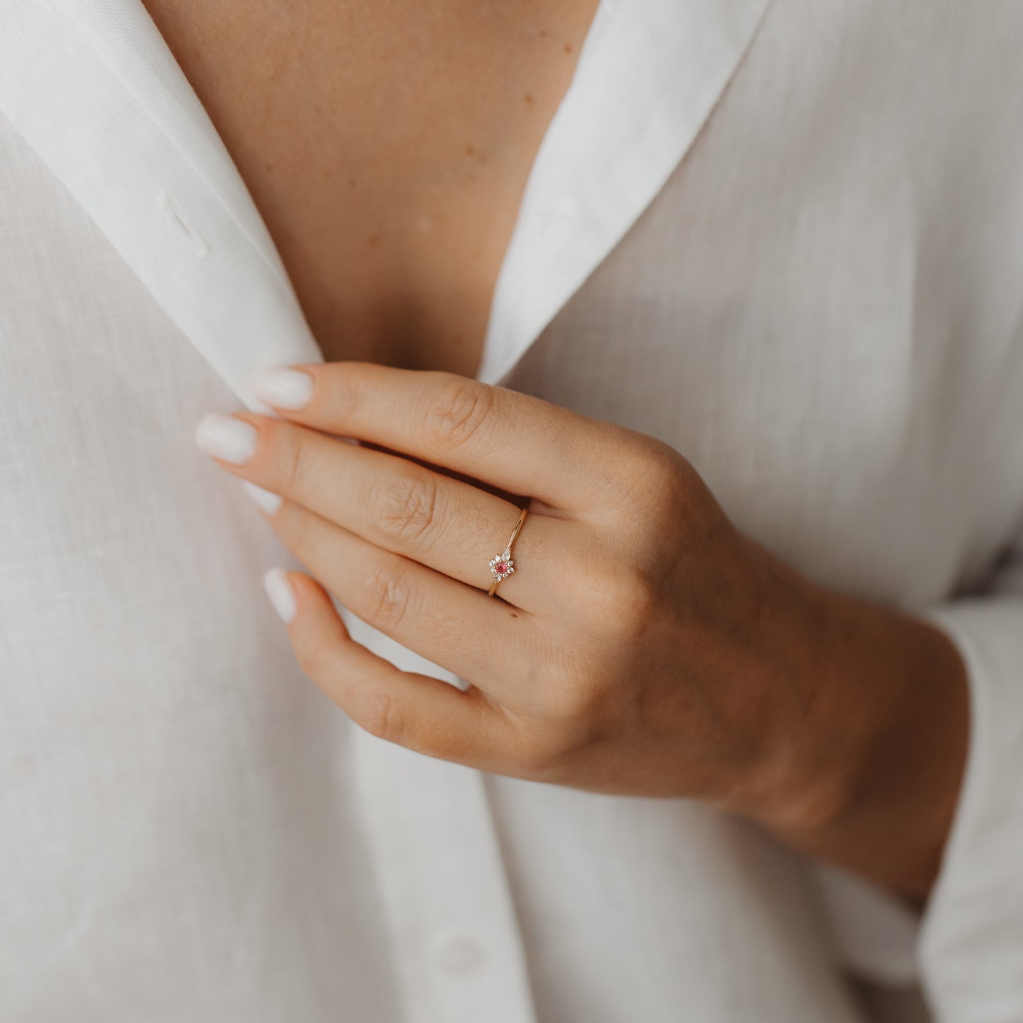 Primo piano di una mano che indossa l'anello Flora in oro giallo 14K con zaffiro bianco e tormalina rosa a grappolo floreale, sullo sfondo di una camicia di lino bianca.
