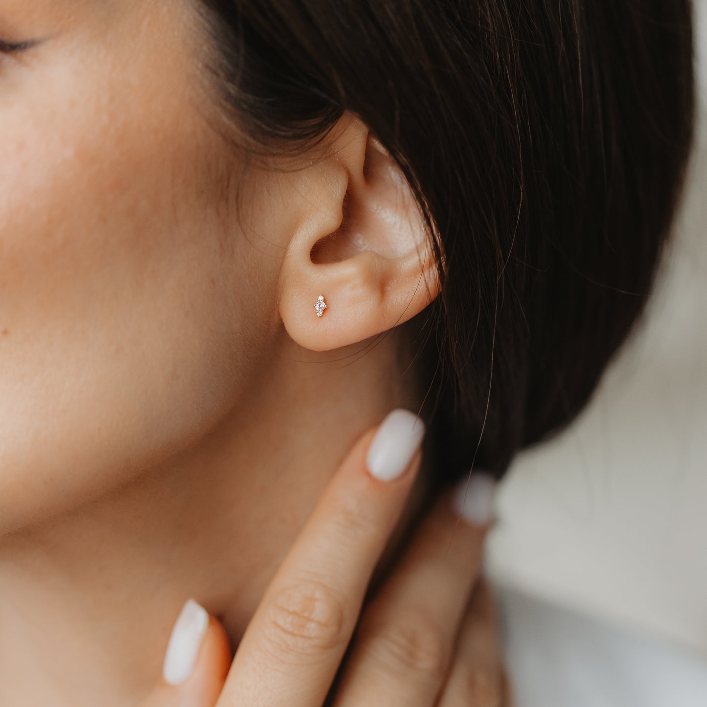 Primo piano di un orecchio che indossa un piccolo orecchino a perno Flora in oro giallo 14K con morganite rosa e zaffiro bianco, mentre una mano con manicure neutra tocca il collo.