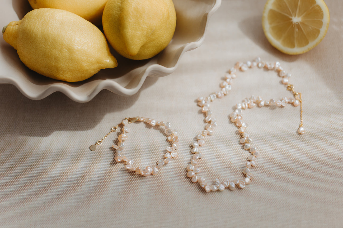 Collana di perle keshi rosa e bracciale coordinato in argento placcato oro, posati su lino beige con limoni freschi.