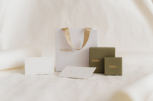 Foto in studio minimalista ed elegante del set di confezioni regalo INDIRA: una borsa bianca con manici in nastro dorato, due scatole per gioielli color oliva e biglietti vuoti su uno sfondo avorio tenue.