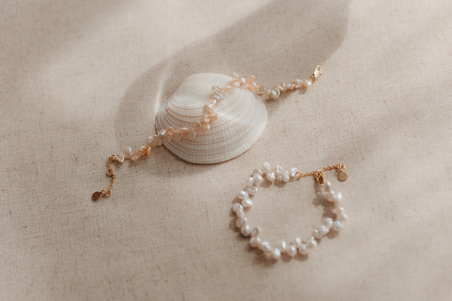 Due bracciali in argento sterling placcati in oro con perle keshi rosa, adagiati su una conchiglia su tessuto di lino beige.