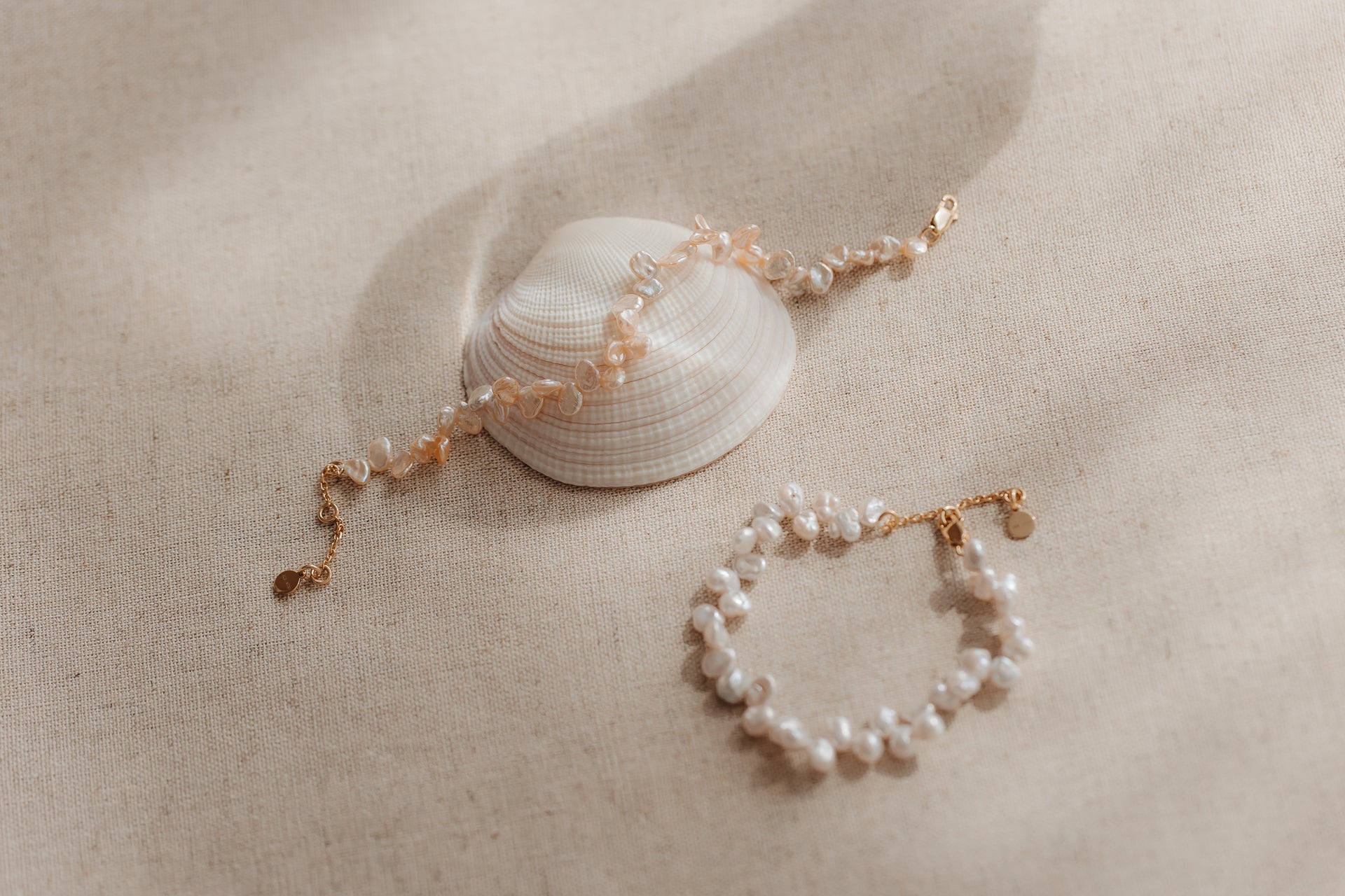 Due bracciali in argento sterling placcati in oro con perle keshi rosa, adagiati su una conchiglia su tessuto di lino beige.