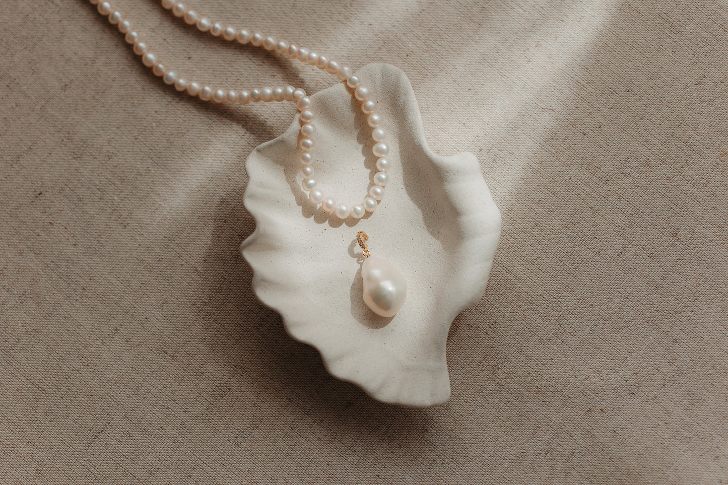 Ciondolo con perla barocca con anello di sospensione in argento placcato oro, appoggiato su un piattino in ceramica a forma di conchiglia con bordo ondulato, con un filo di perle su sfondo di lino beige.