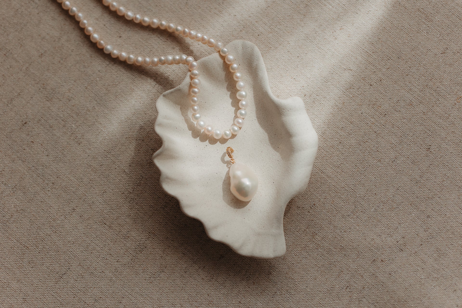 Ciondolo con perla barocca con anello di sospensione in argento placcato oro, appoggiato su un piattino in ceramica a forma di conchiglia con bordo ondulato, con un filo di perle su sfondo di lino beige.