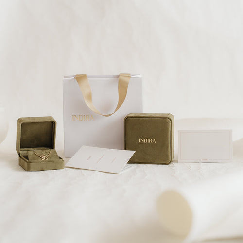 Set di confezioni regalo a marchio Indira con borsa bianca e manici in nastro dorato, eleganti cofanetti portagioie in velluto color oliva (uno aperto) e cartoncini disposti su uno sfondo neutro e morbido.