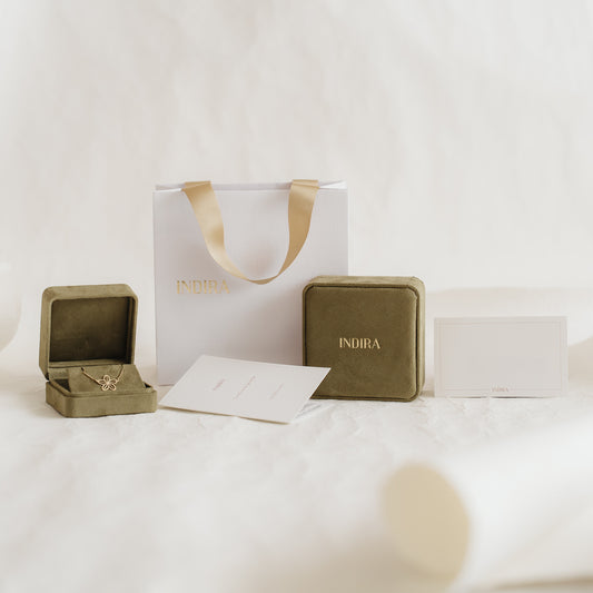 Set di confezioni regalo a marchio Indira con borsa bianca e manici in nastro dorato, eleganti cofanetti portagioie in velluto color oliva (uno aperto) e cartoncini disposti su uno sfondo neutro e morbido.