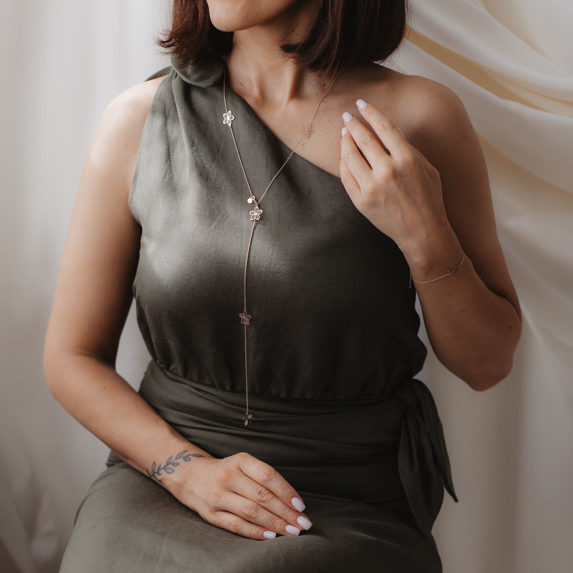 Primo piano di una modella che indossa la collana lariat Wildflower Maxi in oro giallo 14K con charm floreali sopra un abito monospalla color oliva.