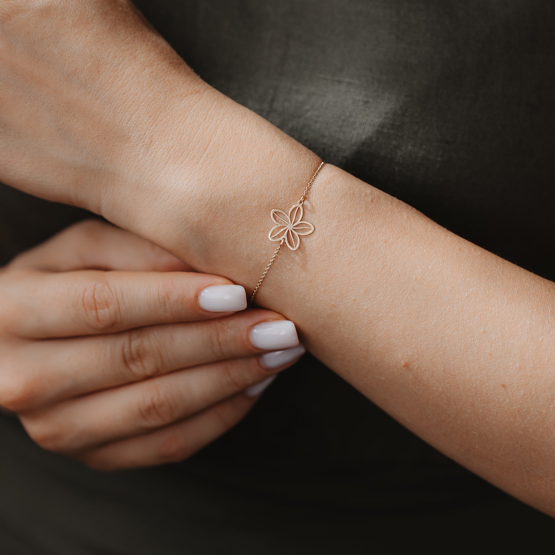 Primo piano di un delicato bracciale in oro 14K con ciondolo traforato a fiore di campo su un polso, su sfondo di tessuto scuro.
