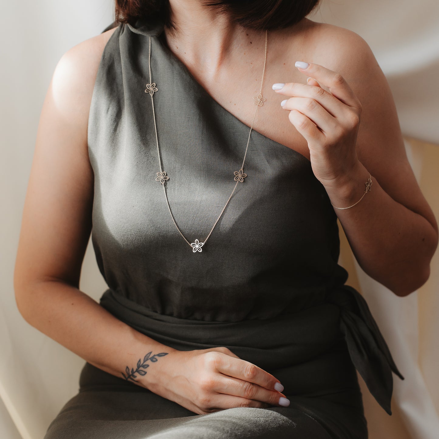 Primo piano di una modella in abito monospalla color oliva che indossa la collana station Wildflower Maxi in oro giallo 14K con piccoli ciondoli a forma di fiore, in una luce interna soffusa, abbinata a un bracciale coordinato.