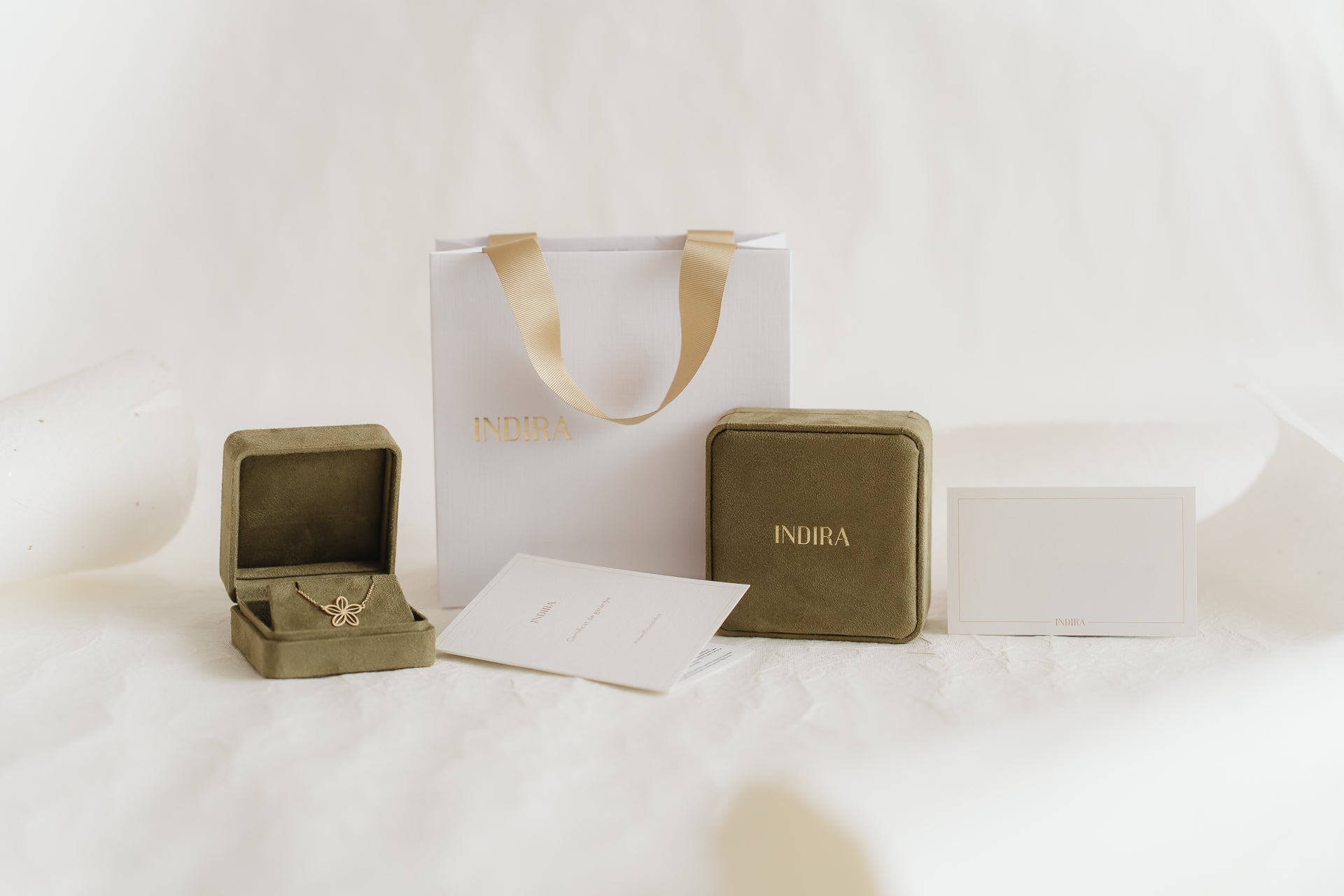 Set per confezione regalo Indira: shopper bianca con manici in nastro dorato, due cofanetti portagioielli in velluto color oliva (uno aperto che mostra un pendente a forma di fiore in oro) e cartoncini brandizzati su un morbido sfondo bianco da studio.